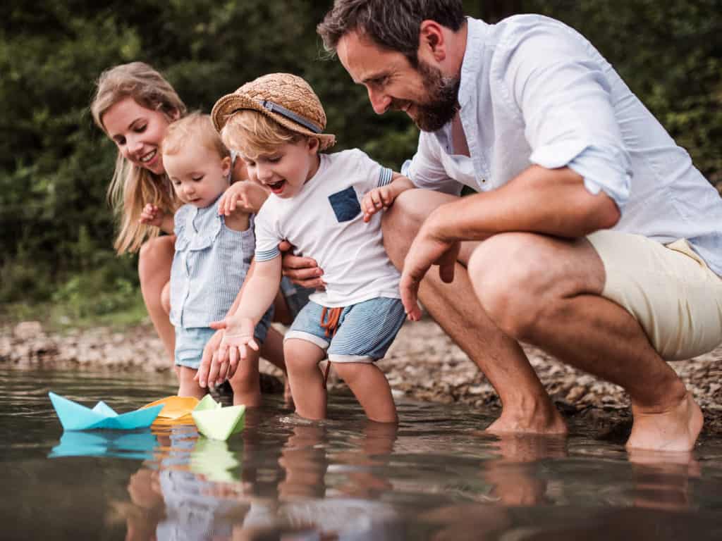 Young family with two toddler children outdoors by the river in summer, playing.