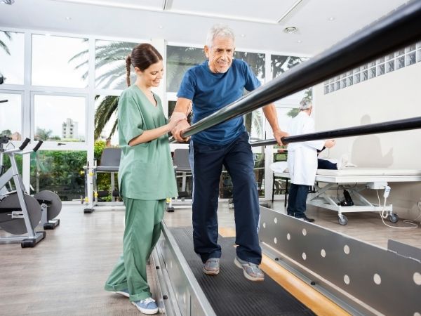 Physiotherapist assisting an older male patient walking on a treadmill in a rehabilitation clinic with gym equipment and natural lighting in the background.