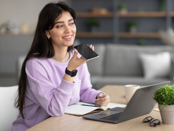 Young woman using voice search on her mobile device while working on a laptop at home