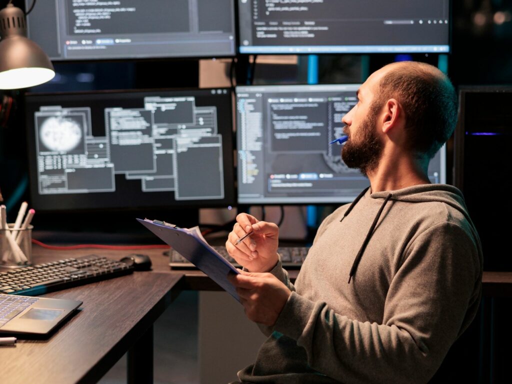 Jason - Website developer sitting at a desk with multiple screens displaying code, appearing disengaged while reviewing notes in a tech workspace